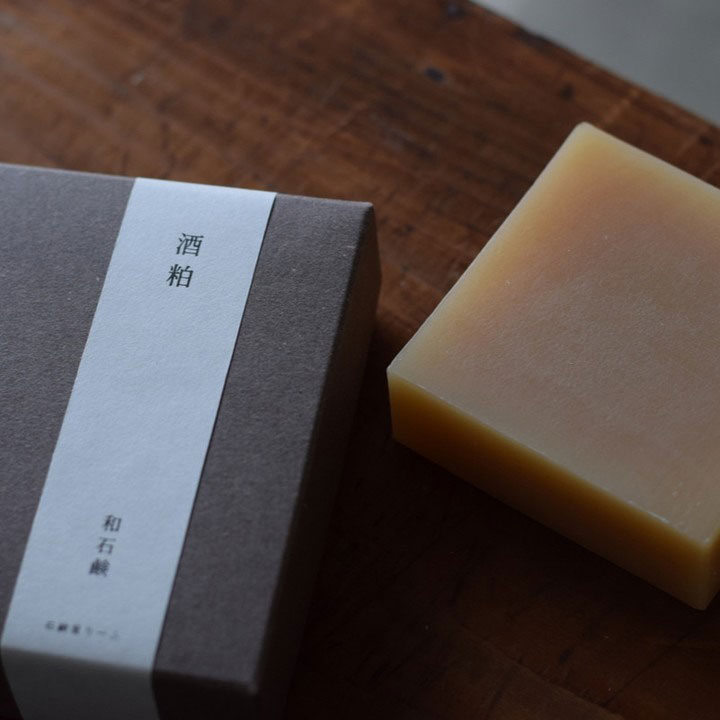 茶色の箱に「酒粕」と印字された帯が巻かれ、琥珀色の石鹸が木のテーブルに並んでいる様子